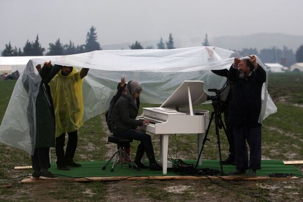 الفنانة الصينية آي ويوي (يمين الصورة) تساعد في حمل واق مم المطر فوق شابة سورية وهي تعزف على البيانو على الحدود اليونانية المقدونية بالقرب من قرية إيدوميني اليونانية، حيث حوصر الآلاف من اللاجئين والمهاجرين بسبب الحصار على حدود البلقان، في مارس 2016 - سبوتنيك عربي