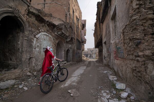 امرأة عراقية ترتدي زي بابا نويل تنظر إلى المباني المدمرة وهي تمشي بدراجتها وسط انتشار مرض فيروس كورونا (كوفيد-19)، في مدينة الموصل القديمة، العراق 18 ديسمبر 2020 - سبوتنيك عربي