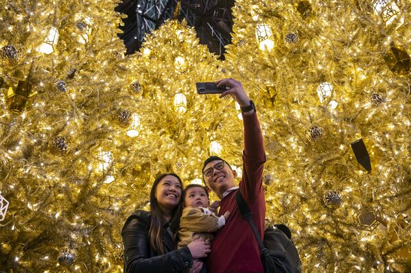 زوجان مع طفلهما يلتقطان صورة سلفي على خلفية أشجار عيد الميلاد الذهبية في أحد مراكز التسوق التجارية في هونغ كونغ، 19 ديسمبر 2020 - سبوتنيك عربي