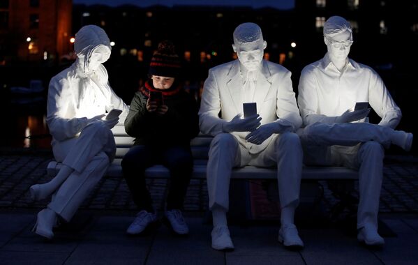 صبي يجلس على تمثال على شكل مقعدعليه أشخاص ينظرون إلى هواتفهم المحمولة، وهو يجلس بجوار جزء من تمثال مضاء الممتص من قبل الإضاءة، للفنان غالي ماي لوكاس، في ليفربول، بريطانيا، 22 مارس 2021 - سبوتنيك عربي