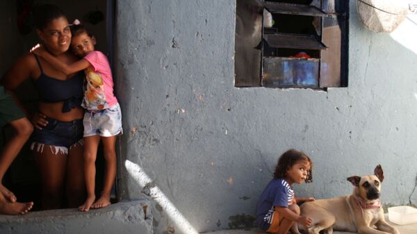 مونيك فيرناندا دي أسيس، 26 عامًا، تقف عند باب منزلها مع ابنتها إستير، حيث يلعب ابنها ميغيل مع كلبهم، بعد تلقي الطعام المتبرع به من مشروع Uma Mao Lava a Outra (يد واحدة تغسل الأخرى) خلال جائحة كورونا (كوفيد-19) في نيلوبوليس، ولاية ريو دي جانيرو، البرازيل، 6 أبريل 2021 - سبوتنيك عربي