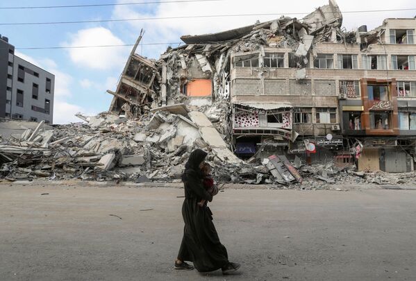 امرأة تحمل طفلها تسير على خلفية ركام منازل، تخلو حييها بعد اشتداد قصف المدافع والطيران الحربي الإسرائيلي في شمال قطاع غزة، فلسطين 14 مايو 2021 - سبوتنيك عربي