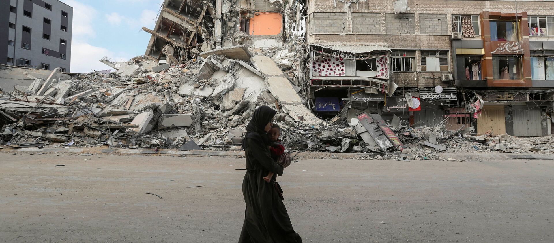 امرأة تحمل طفلها تسير على خلفية ركام منازل، تخلو حييها بعد اشتداد قصف المدافع والطيران الحربي الإسرائيلي في شمال قطاع غزة، فلسطين 14 مايو 2021 - سبوتنيك عربي, 1920, 14.05.2021
