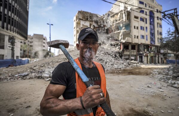 عامل فلسطيني يدخن ويخرج الدخان من أنفه، بينما يأخذ استراحة على خلفية مبنى مدمر، بعد تبادل إطلاق النار بين قطاع غزة وإسرائيل في مايو الماضي، 5 يونيو 2021 - سبوتنيك عربي
