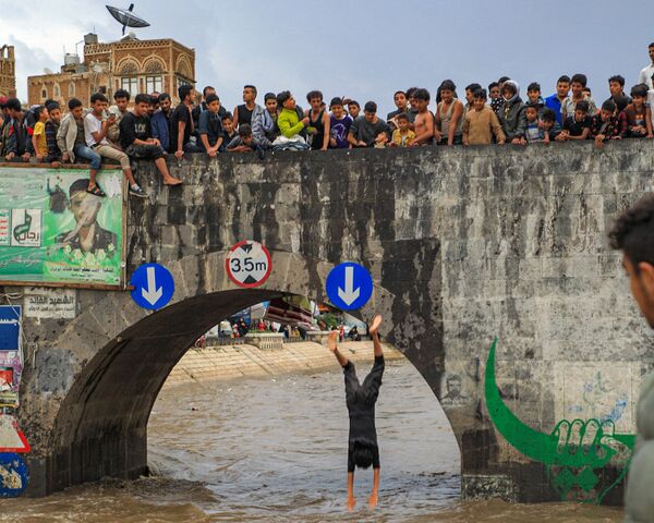 شبان يشاهدون أحدهم يقفز من جسر عبر قناة تصريف غمرتها المياه بعد هطول أمطار غزيرة في المدينة القديمة صنعاء، اليمن في 3 أغسطس 2021 - سبوتنيك عربي