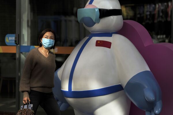 امرأة ترتدي كمامة تسير أمام شخصية تصور موظفاً طبيًا يحارب فيروس كورونا، معروضًا في مبنى مكتب تجاري في بكين، 26 أكتوبر 2021. - سبوتنيك عربي