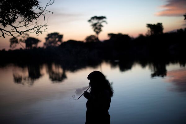 امرأة من قبيلة کامایورا تدخن الأعشاب طقوس صيد الأسماك ضمن طقوس كواروب (Kuarup)، وهي طقوس جنائزية لتكريم ذكرى Cacique Aritana ، زعيم ياوالابيتي، في حديقة شينغو الوطنية، البرازيل، 5 سبتمبر 2021. - سبوتنيك عربي
