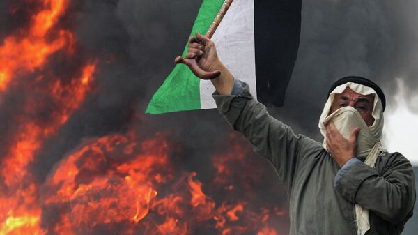 متظاهر فلسطيني يرفع علم فلسطين أمام حاجز من الإطارات المشتعلة في البرقة، الضفة الغربية المحتلة، 23 ديسمبر 2021 - سبوتنيك عربي