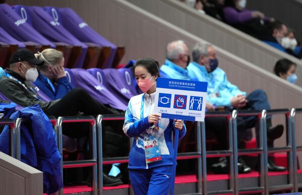 متطوعة تحمل لافتة للحفاظ على المسافة وارتداء الكمامات في الملعب الذي يستضيف منافسة الفرق في التزلج على الجليد في دورة الألعاب الأولمبية الشتوية الـ24 في بكين 2022. - سبوتنيك عربي