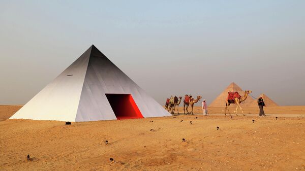 Погонщик верблюдов ведет свое стадо мимо фотобудки в форме пирамиды французского художника JR на плато Пирамид в Гизе - سبوتنيك عربي