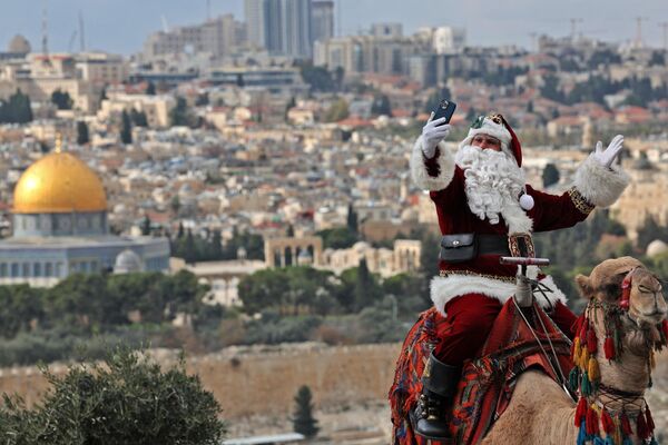 الفلسطيني عيسى قسيسية، مرتديًا زي بابا نويل، يقف لالتقاط صورة بينما يركب جملًا في جبل الزيتون في القدس، قبل أسابيع قليلة من عطلة عيد الميلاد القادمة، 6 ديسمبر 2022. - سبوتنيك عربي