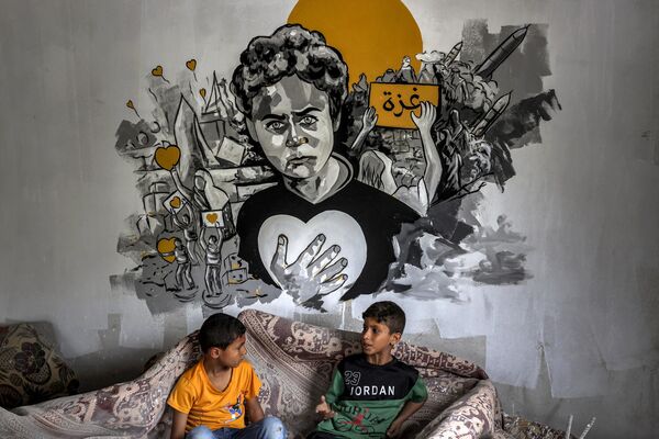 أطفال فلسطينيون يجلسون تحت جدارية خلال معرض فني مختلط بعنوان &quot;الاحتلال يقتل الطفولة&quot;، أقيم وسط أنقاض المنازل التي دمرتها الغارات الجوية الإسرائيلية خلال التصعيد العسكري الأخير في مايو/ أيار 2023، في دير البلح وسط قطاع غزة في 8 يونيو/حزيران 2023. - سبوتنيك عربي