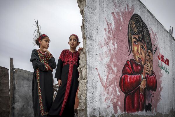 أطفال فلسطينيون يقفون أمام لوحة جدارية خلال معرض فني مختلط بعنوان &quot;الاحتلال يقتل الطفولة&quot; يقام وسط أنقاض المنازل التي دمرتها الغارات الجوية الإسرائيلية خلال التصعيد العسكري الأخير في مايو 2023 في دير البلح وسط قطاع غزة، 8 يونيو 2023. - سبوتنيك عربي