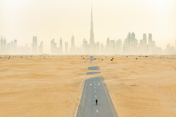 منظر من الأعلى، صورة جوية مذهلة لشخص يمشي على طريق مهجور مغطى بالكثبان الرملية على خلفية مدينة دبي، الإمارات العربية المتحدة - سبوتنيك عربي