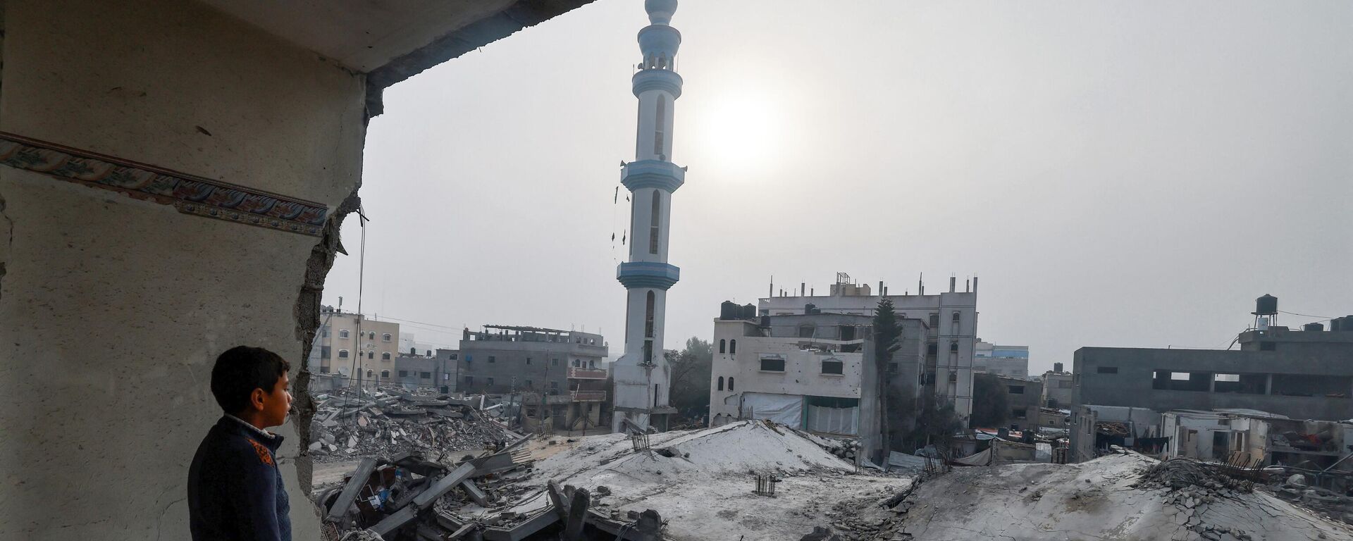 طفل يقف داخل مبنى متضرر ويحدق  بمسجد الفاروق، الذي دمره القصف الإسرائيلي في رفح بجنوب قطاع غزة. - سبوتنيك عربي, 1920, 06.03.2024
