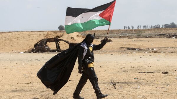 شاب فلسطيني يحمل العلم الفلسطيني، مرتديا زي ارجل الوطواط، خلال فعاليات مسيرات العودة في شرق خانيونس، على الحدود بين قطاع غزة وإسرائيل، 9 مايو/ أيار 2018 - سبوتنيك عربي