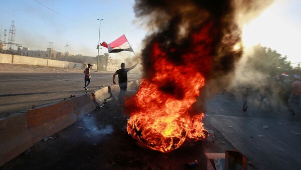 متظاهر يحمل العلم العراقي بالقرب من الأجسام المحترقة في احتجاج أثناء حظر التجول - سبوتنيك عربي
