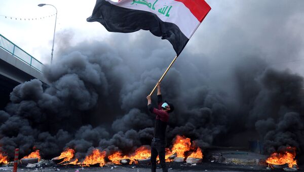 متظاهر يحمل العلم العراقي بجانب الإطارات المحترقة أثناء الاحتجاجات المستمرة المناهضة للحكومة في كربلاء - سبوتنيك عربي