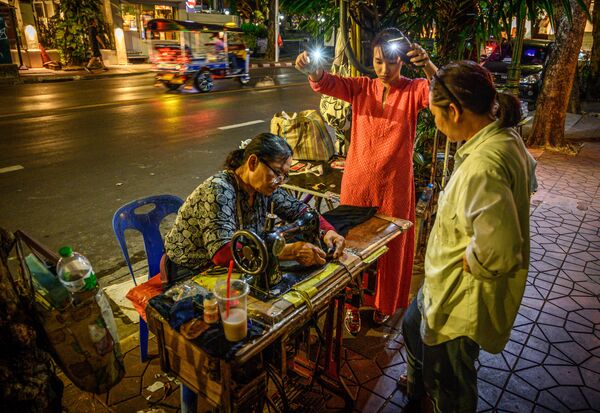 امرأة تستخدم إنارة الهواتف المحمولة لتساعدها أثناء الخياطة في أحد شوارع بانكوك، تايلاند 3 ديسمبر 2019 - سبوتنيك عربي