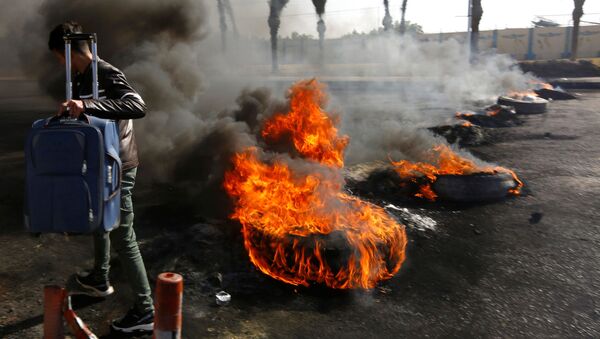 راكب يحمل حقيبته وهو يمشي عبر إطارات محترقة أشعلها المحتجون العراقيون لإغلاق الطريق المؤدي إلى مطار النجف الدولي خلال الاحتجاجات المستمرة المناهضة للحكومة - سبوتنيك عربي