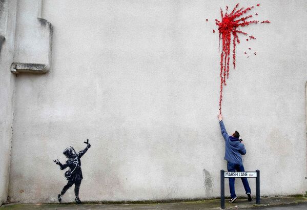 أحد المارة يقف على خلفية رسم غرافيتي لفنان الشارع الشهير بانكي، الرسم لفتاة صغيرة تطلق مقلاعًا من الزهور، على جدار مبنى في بريستول، بريطانيا 14 فبراير 2020. - سبوتنيك عربي