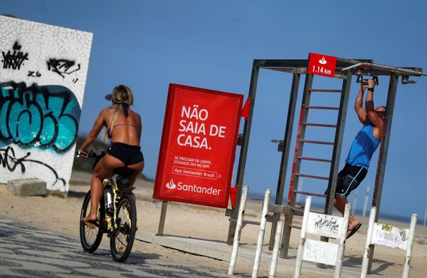 امرأة تركب دراجة تمر بالقرب من لافتة كتب عليها لا تغادر المنزل على شاطئ إيبانيما، أثناء تفشي مرض فيروس كورونا (كوفيد-19) في ريو دي جانيرو، البرازيل 5 أبريل 2020 - سبوتنيك عربي