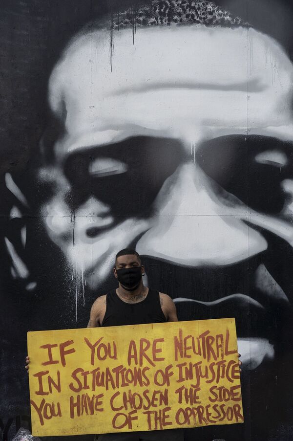 رجل يقف على خلفية جدارية المواطن الأمريكي من أصول أفريقية، جورج فلويد، ويحمل لافتة كتب عليها (إذا كنت محايداً في قضية ظالمة، فأنت إخترت جانب المضطهد) في مينيابوليس، بولاية مينيسوتا،2 يونيو 2020 - سبوتنيك عربي
